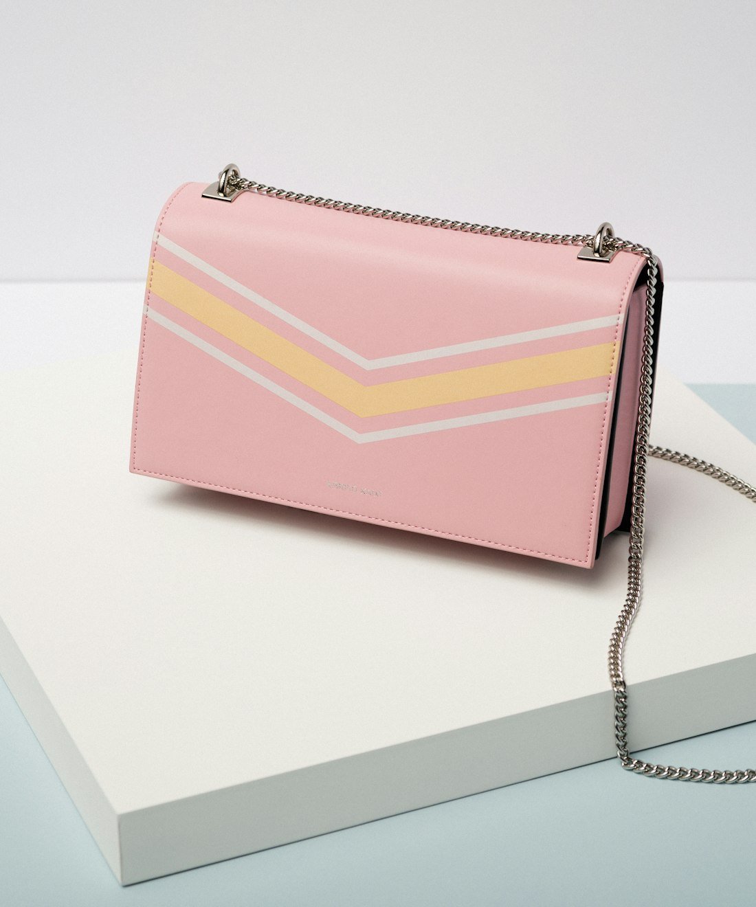 Blush pink flap handbag with a slim silver chain strap, photographed on a pale display block